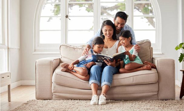 Young happy family sitting on the sofa and reading a book together at home while relaxing. a young couple reading a book to their daughters while sitting on a sofa at home - Foto, Bild