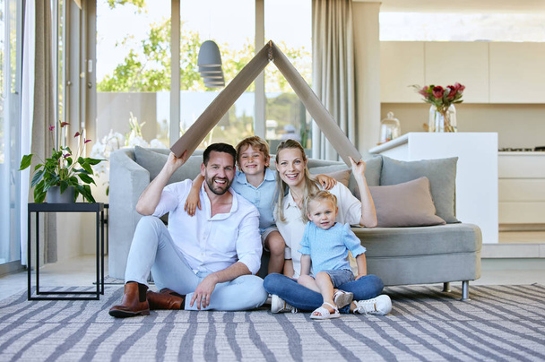 Full length portrait of an affectionate family of four holding a cardboard roof over their heads while sitting in the living room at home. - Foto, Bild