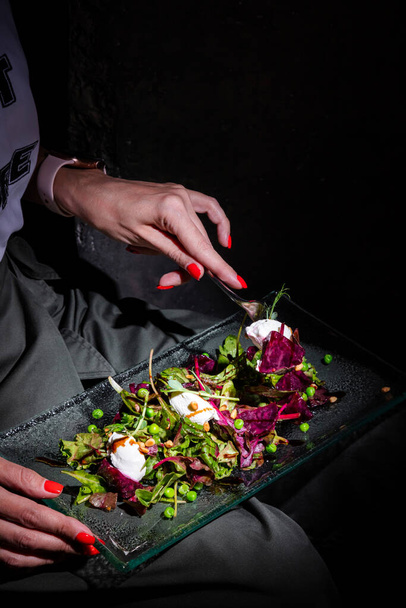 Man holds a glass plate in his lap and eats with a fork salad with mozzarella, arugula, seeds, peas, beets, frisse salad and sauce - Foto, Imagen