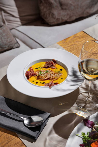 Pumpkin porridge with bread, seeds, prosciutto and pea sprouts in a plate on the table with glasses of wine, cutlery, a tablecloth and a napkin on the background - Fotó, kép