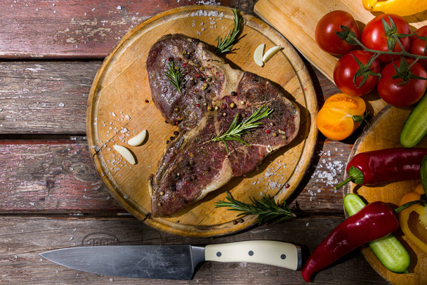 Grilled beef meat on the bone with cloves of garlic, tomatoes, red hot and sweet peppers, cucumbers, rosemary, salt and peppercorns with a knife on a wooden board and a wooden table. Horizontal orientation - Фото, изображение