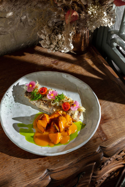 Запечённая рыба с лепестками цветов, жареная картошка в яичном желтке, чука и лук в тарелке на столе - Фото, изображение