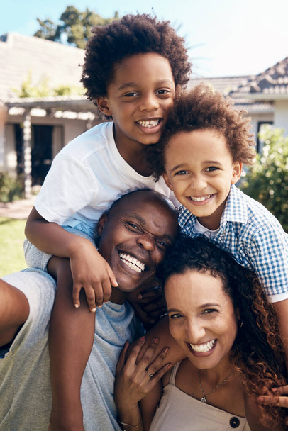 Szczęśliwa afrykańska rodzina czteroosobowa bawiąca się podczas robienia selfie na słońcu. Beztroscy rodzice przewożący synów na przejażdżki na barana podczas nawiązywania więzi na zewnątrz. Mama i tata cieszą się dobrym czasem z dziećmi. - Zdjęcie, obraz