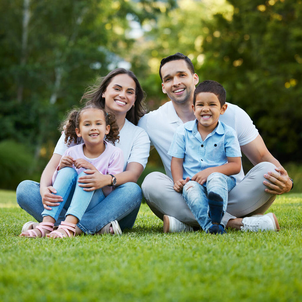 Młoda szczęśliwa mieszana rodzina rasy relaksująca się i siedząca razem na trawie w parku. Kochający rodzice spędzający czas ze swoimi dziećmi w ogrodzie. Beztroska więź rodzeństwa z mamą i tatą. - Zdjęcie, obraz