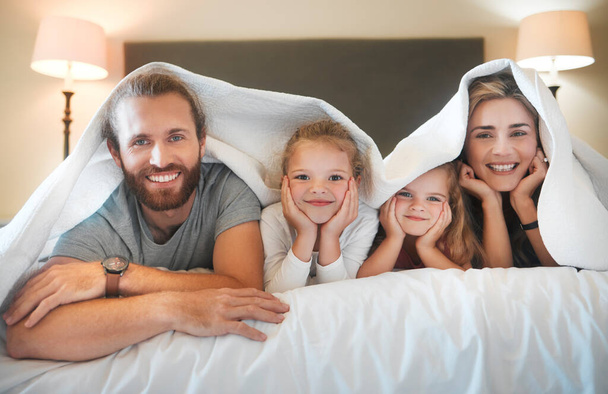 Młoda, kaukaska rodzina leżąca pod kocem w łóżku w domu. Cute little rodzeństwo relaks w łóżku z matką i ojcem w sypialni. - Zdjęcie, obraz