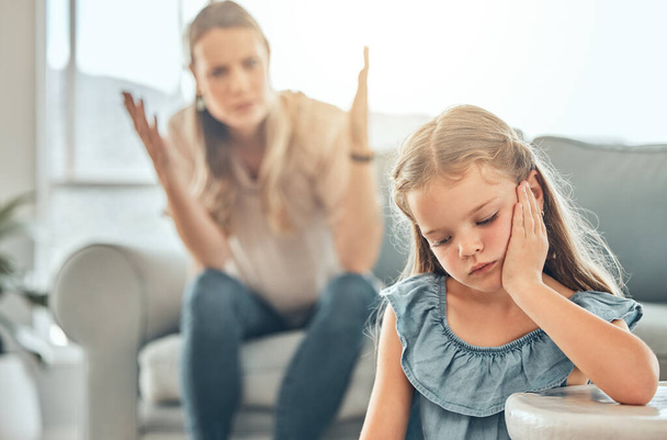 Sad little caucasian girl sitting in the living room and feeling depressed while her mother shouts and raises her hands behind her. Young cute daughter feeling upset after disappointing her parent at. - Foto, afbeelding