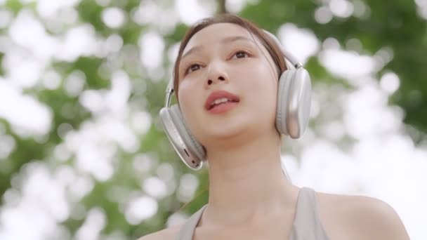 Asian woman is engaged in fitness in public park. Female wearing sport bra and using headphone listening to music. Healthy lifestyle concept, sports outdoor activities in park - Video, Çekim