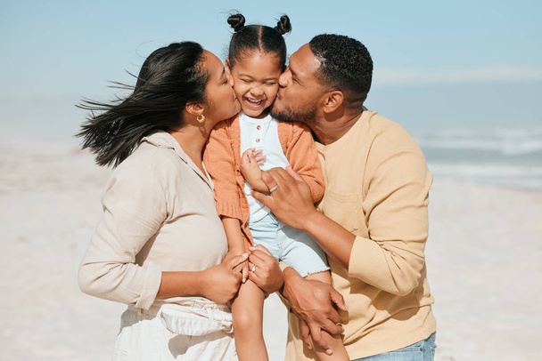 Happy mixed race family standing on the beach. Loving parents kissing adorable little daughter on the cheeks showing love and affection while enjoying beach vacation. - Foto, Imagen