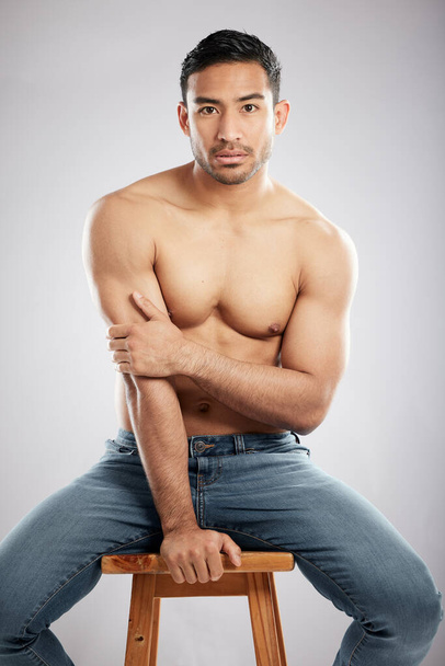 Always facing the right way. Studio shot of a handsome young man showing off his muscular body while sitting on a chair against a grey background - Fotoğraf, Görsel