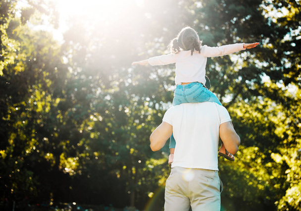 Vader en speelse dochter van achteren hebben plezier in de zon in het park met copyspace op groene bomen. Liefdevolle ouder dragen vrolijk meisje op de schouders voor piggyback rit met armen uit te doen alsof ze vliegen. - Foto, afbeelding