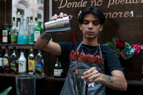 Young latin bartender at a bar pouring cocktails into a shaker. Bartender is making a cocktail. Hispanic bartender working at the bar counter. Bartender mixing alcohol cocktail in cocktail shaker. - Foto, afbeelding