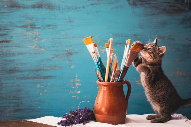 Still life of paint brushes  in clay jug, lavender flowers and a cute kitten on vintage turquoise background - Φωτογραφία, εικόνα