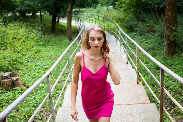 A slender young girl with long blond hair, in a pink dress, walks up the stairs, in the alley of the park, around green trees. He fixes his hair with his hand. - Photo, Image