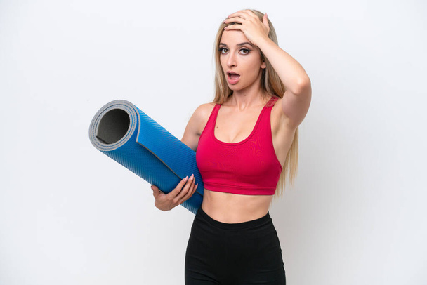 Young sport blonde woman going to yoga classes while holding a mat isolated on white background doing surprise gesture while looking to the side - Photo, Image