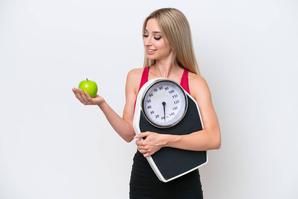 Pretty blonde woman isolated on white background holding a weighing machine while looking an apple - Foto, imagen
