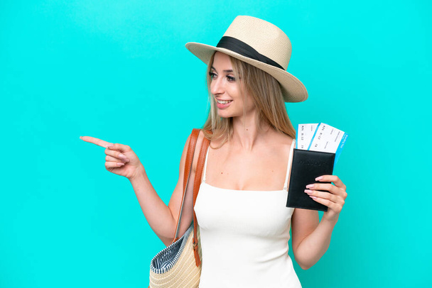 Blonde woman in swimsuit holding passport isolated on blue background pointing to the side to present a product - Фото, зображення