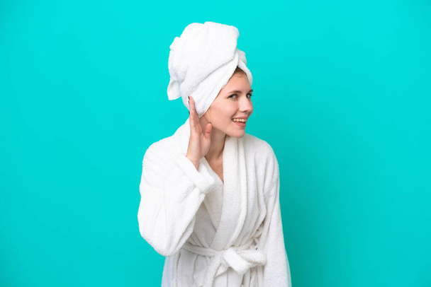 Young woman in a bathrobe isolated on blue background listening to something by putting hand on the ear - Foto, imagen