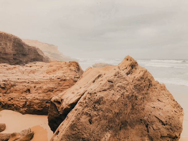 Rocky cliffs on a beach over a background of a cloudy sky - Фото, изображение