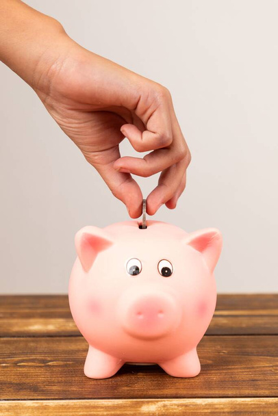 A vertical shot of a hand putting a coin in a piggy bank on a wooden table - Foto, Imagen