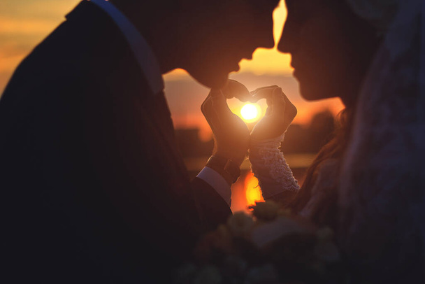 sziluett fiatal házaspár csinál a szív alakját, a napfény áthalad a kezét nézi egymást. naplemente. Az esküvő napján. Valentin napi szerelem koncepció. Koncentrálj a kezekre. - Fotó, kép