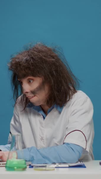 Vertical video: Insane foolish female chemist using laptop and beaker in front of camera, acting crazy and amusing. Mad funny woman with messy wacky hair looking goofy and funny, being silly in studio - Imágenes, Vídeo
