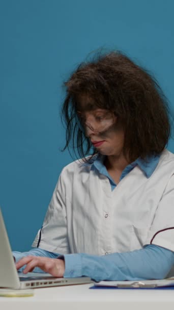 Vertical video: Portrait of crazy female chemist working on laptop at desk, being funny and goofy in studio. Mad insane scientist using computer and doing foolish silly facial expressions, having - Imágenes, Vídeo