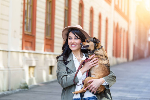 Nuori hymyilevä nainen hattu rento vaatteet Silitysrauta ja halaus koira taas kävelemässä kadulla. Hymyilevä nuori nainen nauttii hyvästä päivästä ja poseeraa lemmikin kanssa. Nainen leikkii koiransa kanssa - Valokuva, kuva