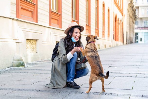 Young woman with long hair and face protective mask embracing funny dog on the street during coronavirus pandemic isolation. Smiling young woman enjoying good day and posing with pet. - Photo, image