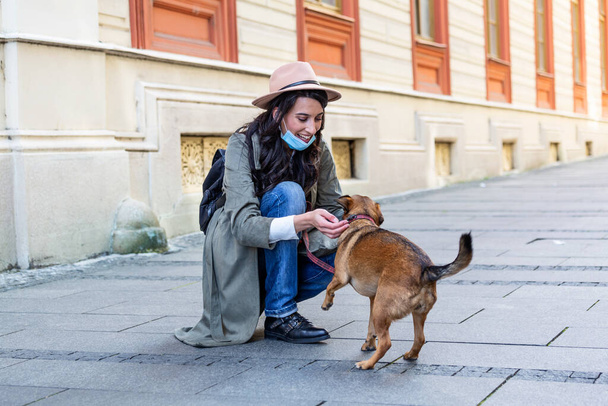 Mulher jovem com cabelos longos e máscara protetora rosto abraçando cão engraçado na rua durante o isolamento pandêmico coronavírus. Sorrindo jovem mulher desfrutando de bom dia e posando com animal de estimação. - Foto, Imagem