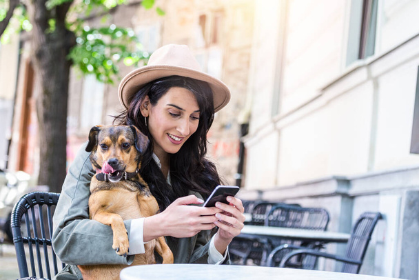 Jonge vrouw met haar hond in huisdiervriendelijke kantine. Jonge vrouw sms 't op haar mobiele telefoon terwijl ontspannen in cafe tijdens vrije tijd met haar hond - Foto, afbeelding