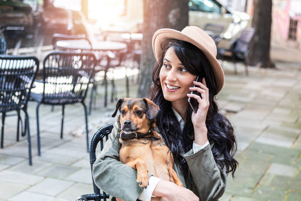 Attractive young woman sitting in coffee shop, relaxing and holding her dog while talking on the phone. Pet friendly cafeteria, beautiful girl with her dog siting at a coffee shop and drink coffee. - Foto, Imagen