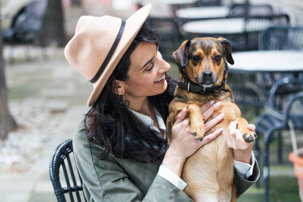 Attractive young woman sitting in coffee shop, relaxing and holding her dog. Pet friendly cafeteria, beautiful girl with her dog siting at a coffee shop and drink coffee. - Valokuva, kuva