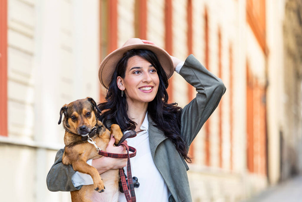 Jonge lachende vrouw met een hoed in casual kleding ijzer en knuffelende hond terwijl ze op straat loopt. Lachende jonge vrouw genieten van een goede dag en poseren met huisdier. Vrouw spelen met haar hond - Foto, afbeelding