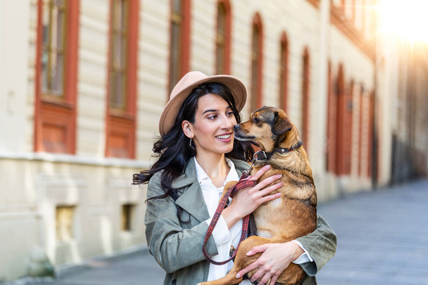 Young smiling woman with a hat in casual clothes iron and hugging dog whilewalking on the street. Smiling young woman enjoying good day and posing with pet. Woman playing with her dog - Foto, Imagen