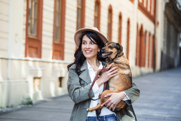 Young smiling woman with a hat in casual clothes iron and hugging dog whilewalking on the street. Smiling young woman enjoying good day and posing with pet. Woman playing with her dog - Zdjęcie, obraz