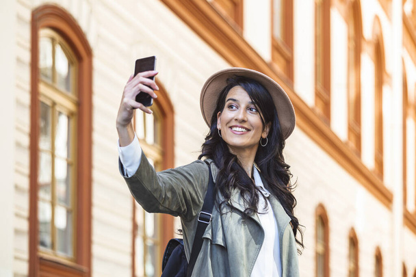 Tourist posing for a selfie in a street. Vlogger recording content for her travel vlog. Cute young smiling girl is making selfie on a camera while walking outdoors. - Zdjęcie, obraz
