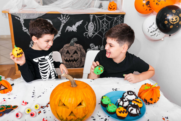 Happy Halloween! Attractive young boy with his brother are preparing for Halloween party. Brothers in costumes are having fun with pumpkins and cupcakes. - Photo, Image