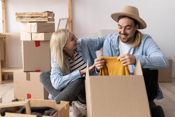 a young cheerful couple in a new apartment on the day of the move sit on the floor unpack things the woman puts on a hat to the man. moving to a new apartment. Lots of packing boxes around them. - Photo, Image