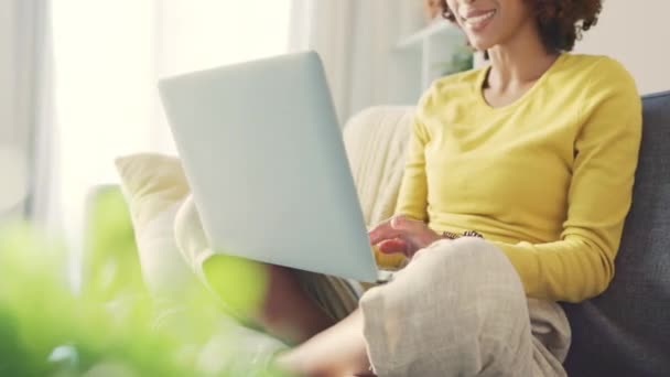 Eine schwarze Frau, die ferngesteuert an einem Laptop arbeitet und zu Hause auf einem Sofa sitzt. Glückliche Studenten tippen und surfen im Internet, während sie recherchieren und planen. Freiberuflicher Unternehmer bloggt online. - Filmmaterial, Video