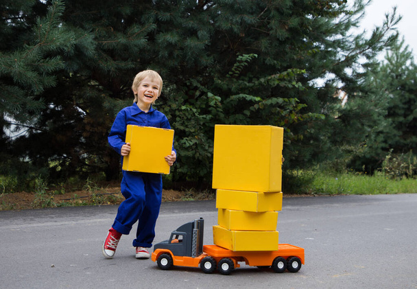 cute smiling boy 5-6 years old in a blue overalls uniform stands near big toy car - truck with a lot of yellow cardboard boxes. Parcel delivery, little postman, Positive, humor, interesting childhood - 写真・画像