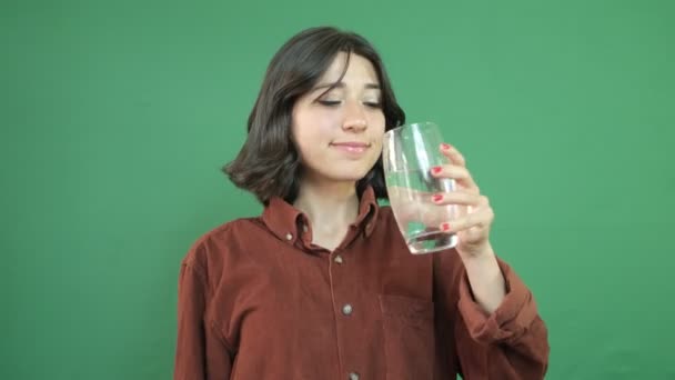 Nainen, joka juo vettä kädessään olevasta lasista, on suhteellinen, juo vettä terveydelle, nuoren naisen ilme vihreän verhon edessä - Materiaali, video