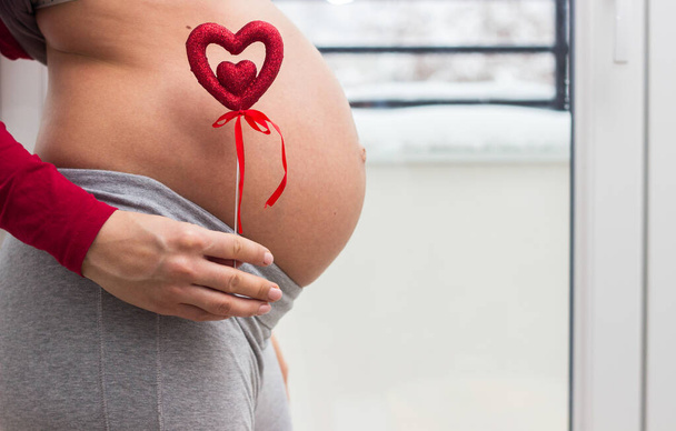 Picture of pregnant woman holding heart sign to one side. Holds hands on belly at home interiors. - Photo, image