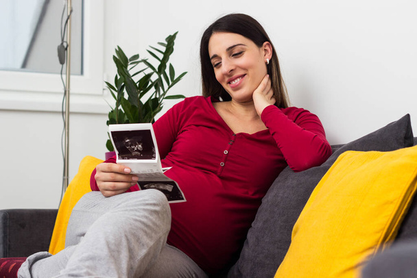 Pregnant woman looking at ultrasound image of her baby. Resting at the home - Zdjęcie, obraz