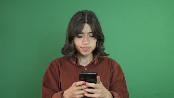 Immagine di una ragazza che messaggia nervosamente al telefono, giovane ragazza che guarda il telefono con un'espressione facciale arrabbiata, espressione facciale di una giovane donna davanti alla tenda verde - Filmati, video