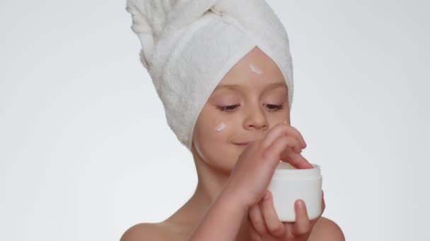 Lovely young child girl with towel on head applying cleansing moisturizing cream from creme jar. Teenager kid face skincare healthy treatment, natural cosmetics. Female portrait. Perfect fresh clean - Záběry, video