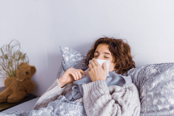 Teenage lying in bed, with warm scarf wrapped around throat, in knitted sweater, sneezes covering mouth with napkin. Colds, sneezing, viral infection, allergic reaction to pets, dust, or houseplants - Photo, image