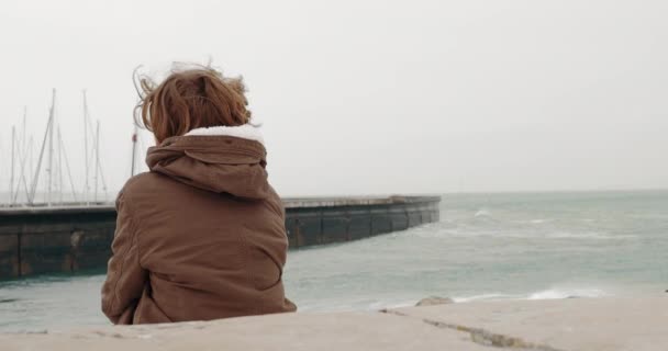 Back view of a boy in warm jacket sitting alone on the coast and watching the ocean waves crushing the breakwater with sailboats parking - Materiał filmowy, wideo