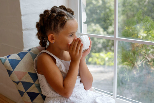 Little girl sitting by the window with a cup of hot drink and looking outdoors - Fotó, kép