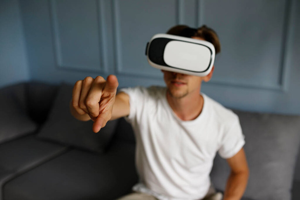 Young man wearing virtual reality goggles in modern coworking interior sitting on sofa. Smartphone using with VR headset. 3D reality concept. - Photo, image