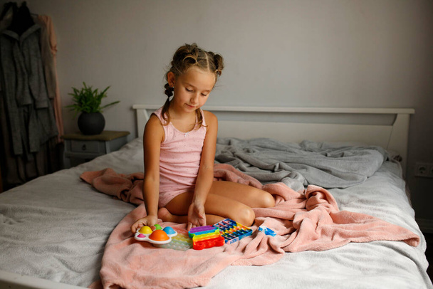 Little cheerful girl plays in a multi-colored plastic toy pop it while lying on bed. Playing at home during summer holidays. Antistress concept for kids - Fotó, kép